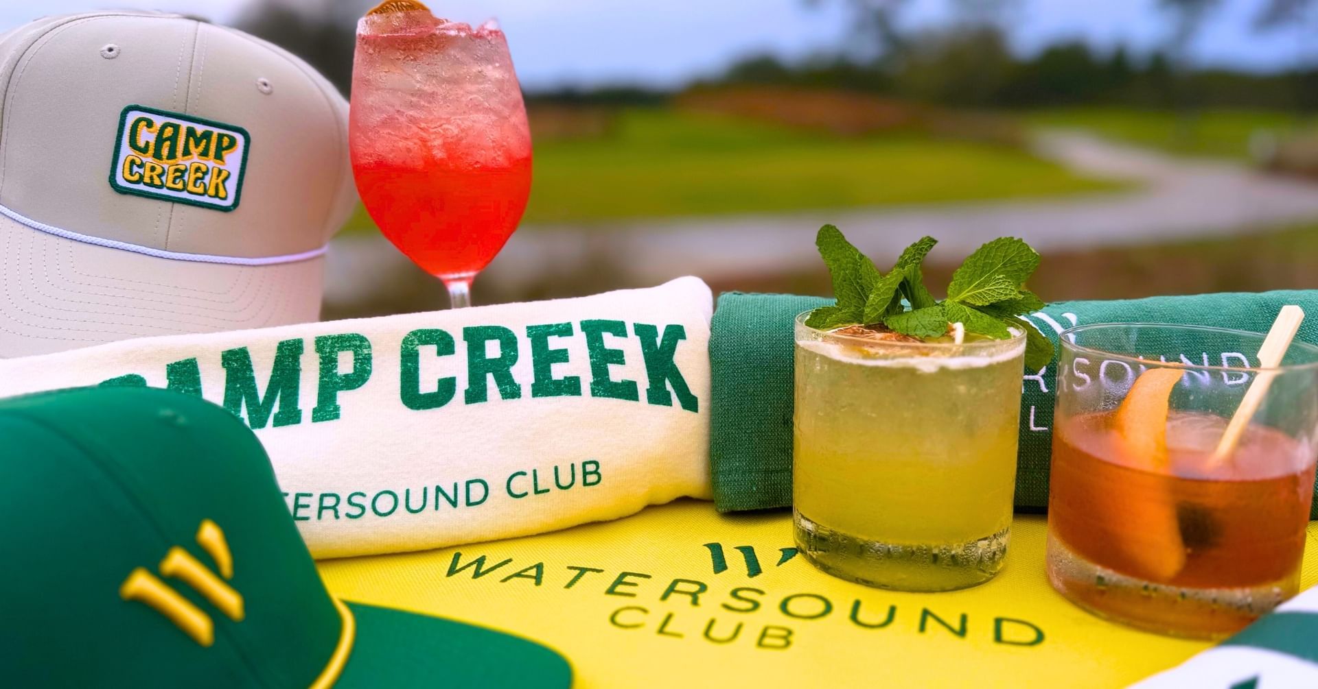Camp Creek branded hats, towel, and cocktails on a yellow surface with
