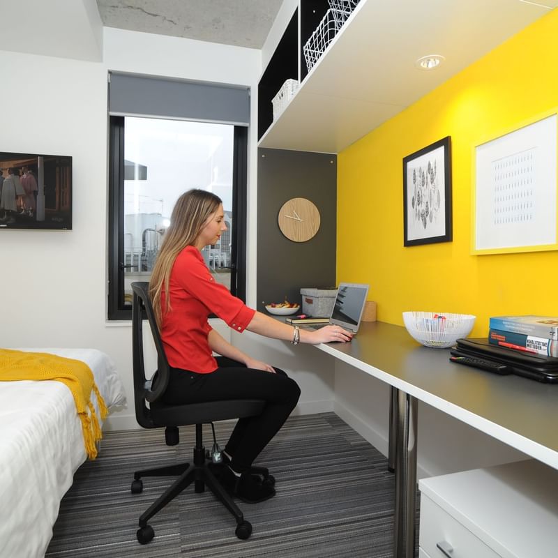 Woman sitting at a desk in a compact living space with a bed at UniLodge Auckland City.