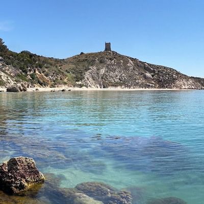 Felsige Küste mit klarem Wasser und Turm auf einem Hügel in der Ferne.