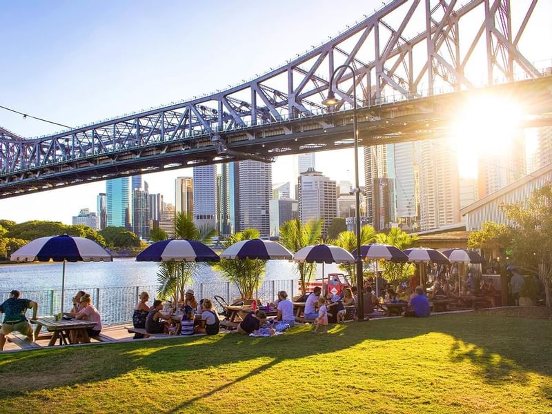 People enjoying a sunny Howard Smith Wharves under umbrellas with a bridge near Sofitel Brisbane Central