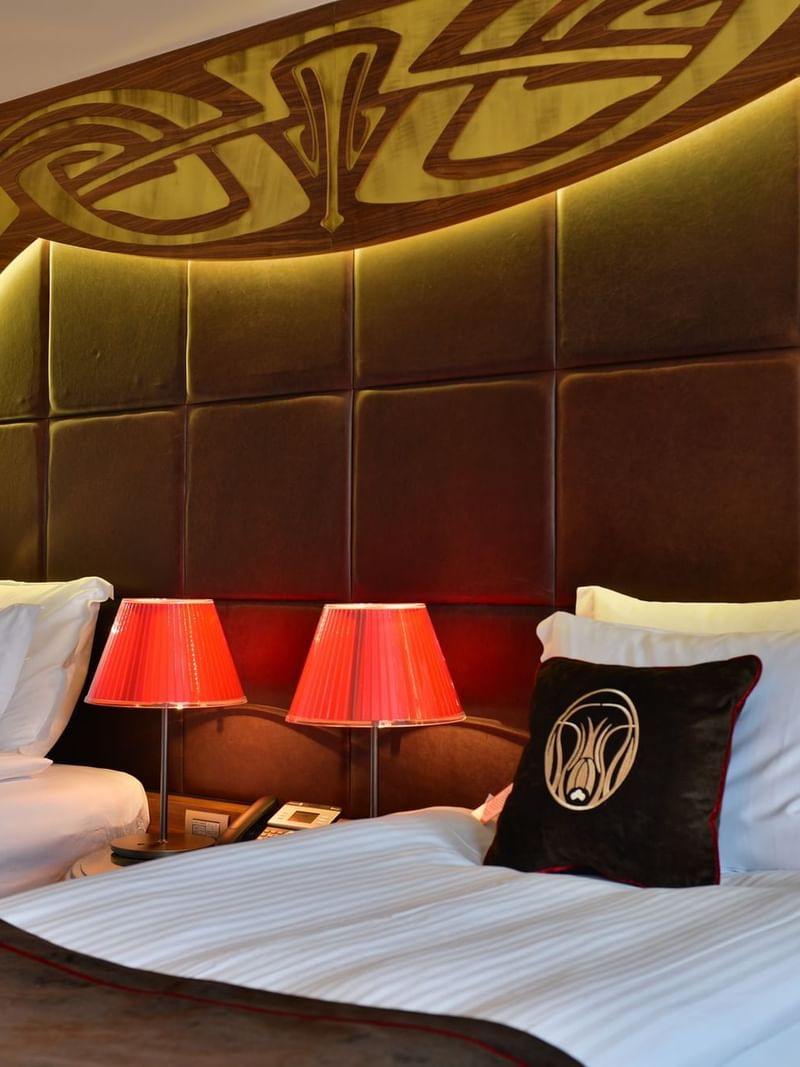 Red lamps by a padded headboard under a wooden arch surrounding twin beds in Deluxe Rooms at Warwick Ankara