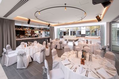 Round tables with white linens by a dark tiled bar under a ring light in the restaurant at the Amora Hotel Group