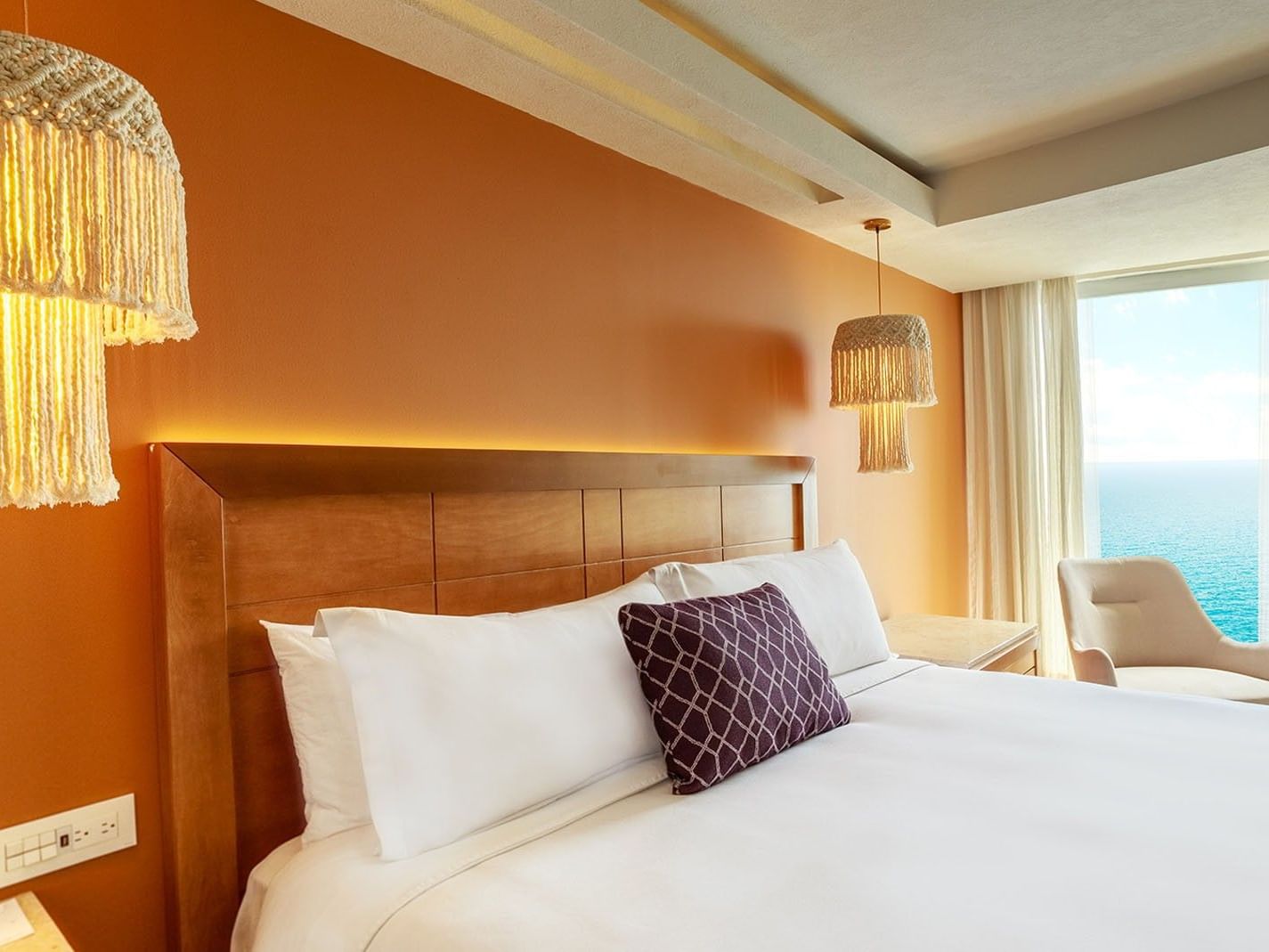 Close-up of a cozy bed in King Deluxe highlighting the warm orange wall and hanging macrame lamps at Camino Real Veracruz
