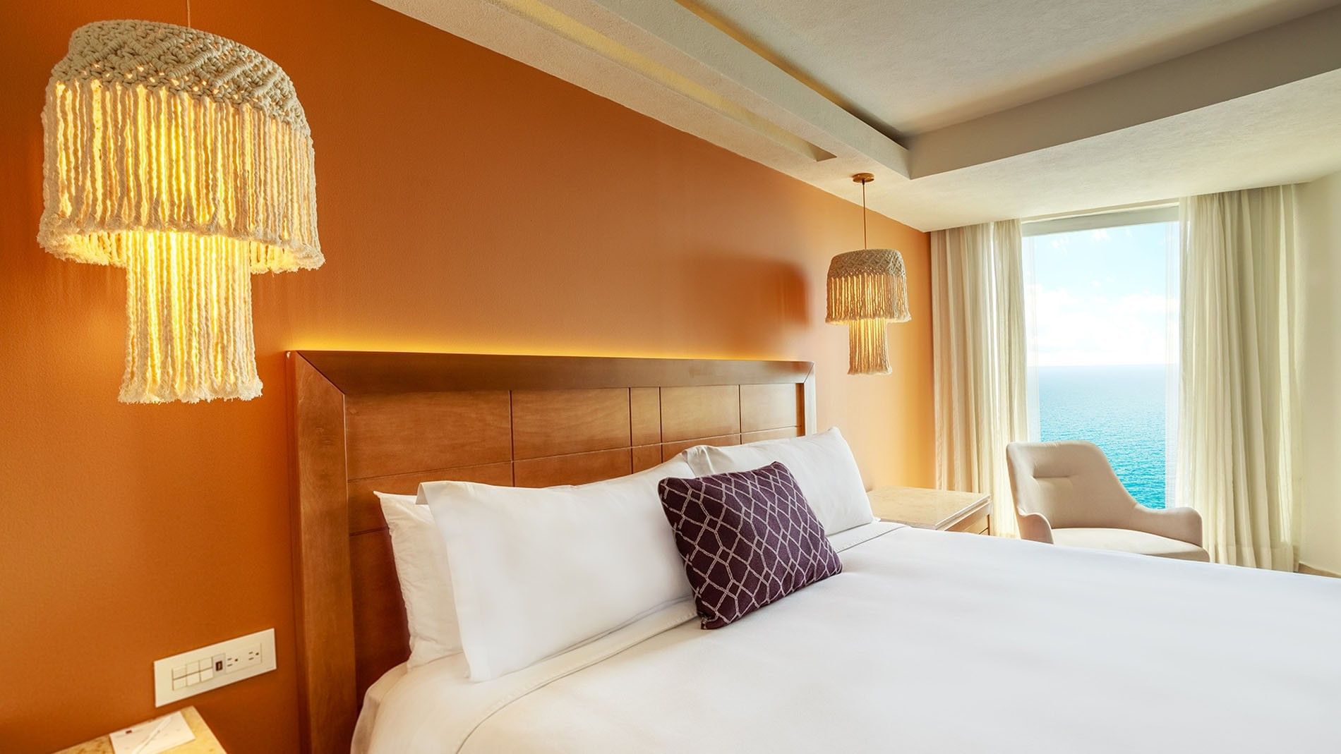 Close-up of a cozy bed in King Deluxe highlighting the warm orange wall and hanging macrame lamps at Camino Real Veracruz