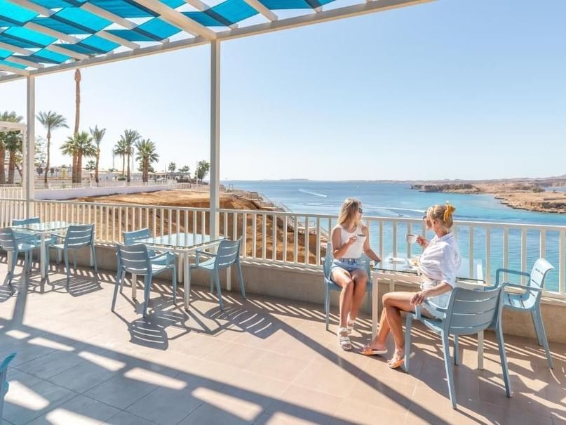 Two women sit at a table on a patio with ocean views, surrounded by blue chairs and tables.