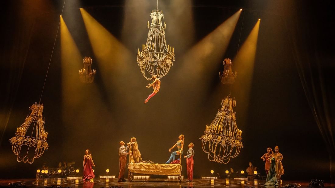 Performers on stage with large chandeliers and spotlights, one person flying mid-air, audience watching below.