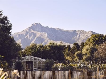 Jardín con valla de madera e invernadero, rodeado de árboles y montañas al fondo.