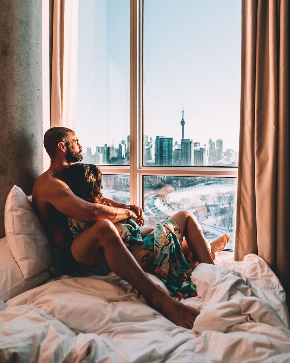 Couple assis sur un lit près de la fenêtre avec vue sur les toits de la ville à l'hôtel X Toronto