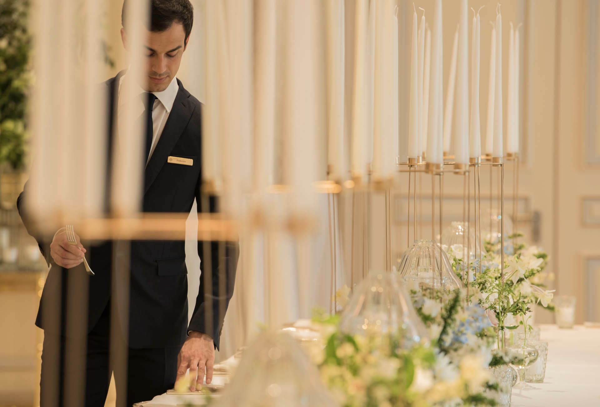 Staff member setting up a table with silverware, floral decor & white pillar candles in an event hall at Palazzo Parigi