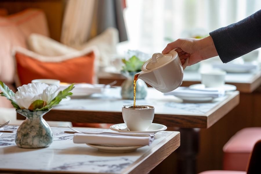 Hand pouring tea into a white cup placed by a floral arrangement at The Capital Hotel, Apartments and Townhouse