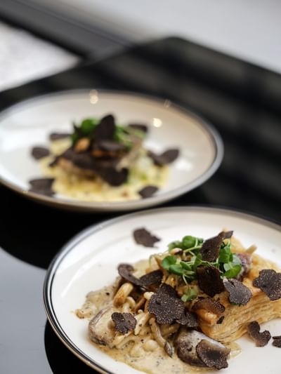 Two elegant plates of gourmet pasta topped with black truffle shavings, mushrooms, and fresh herbs for The Truffle Édition