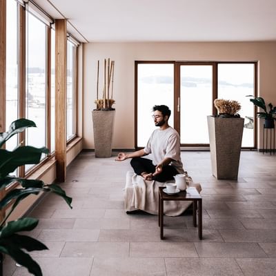 Uomo seduto in meditazione su un cuscino in una stanza luminosa con grandi finestre e piante.