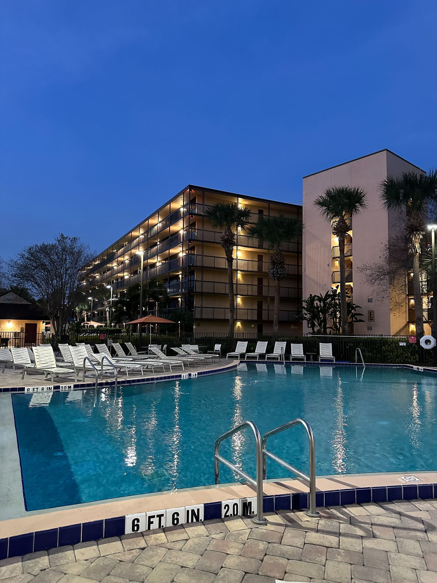 A blue swimming pool reflects the lights of a beige hotel under a dark blue evening sky, with white lounge chairs and palm trees visible around the pool. 