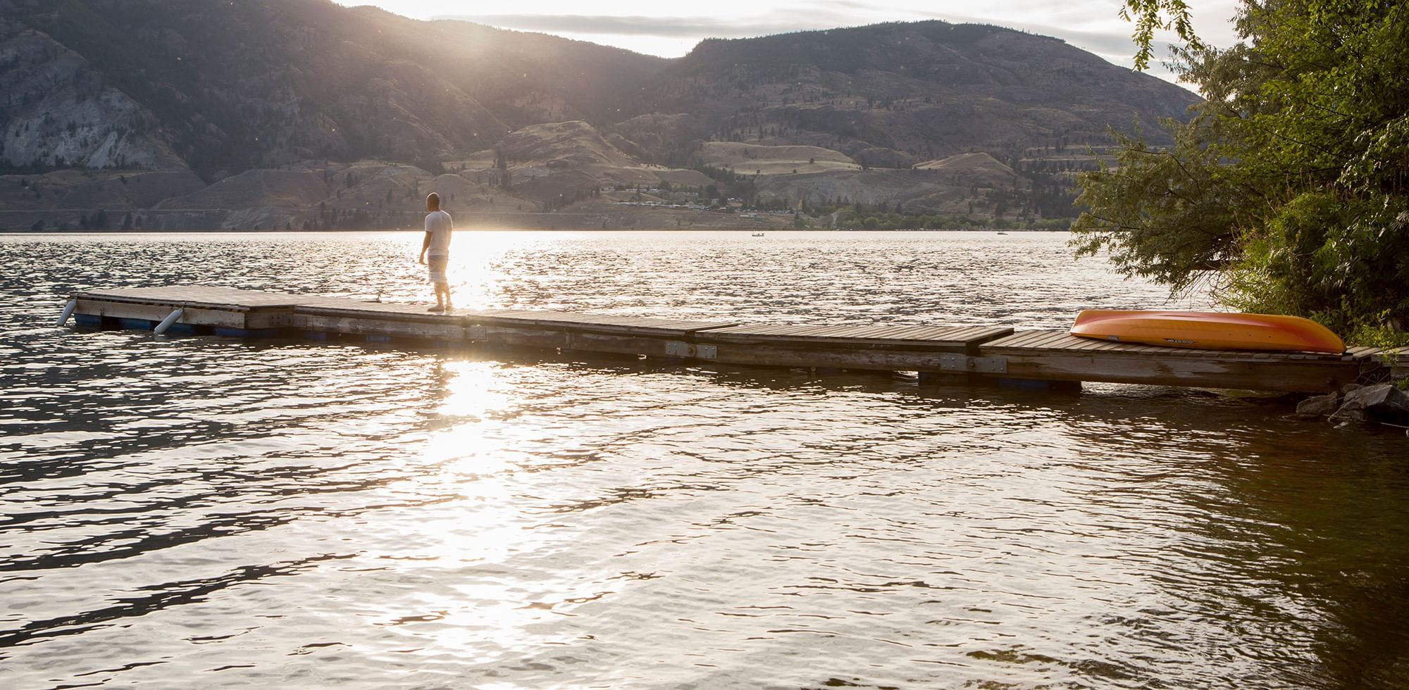 Person standing on a wooden dock over a shimmering lake, with mountains in the background near Coast Penticton Hotel