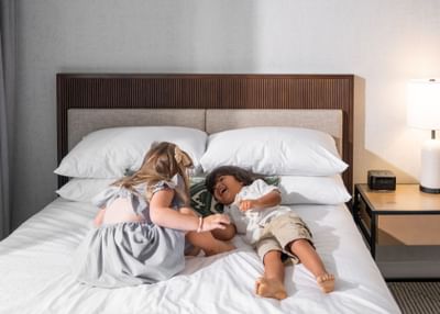 Two kids happily laughing on a bed, surrounded by soft pillows and a wooden headboard at the Maui Coast Hotel