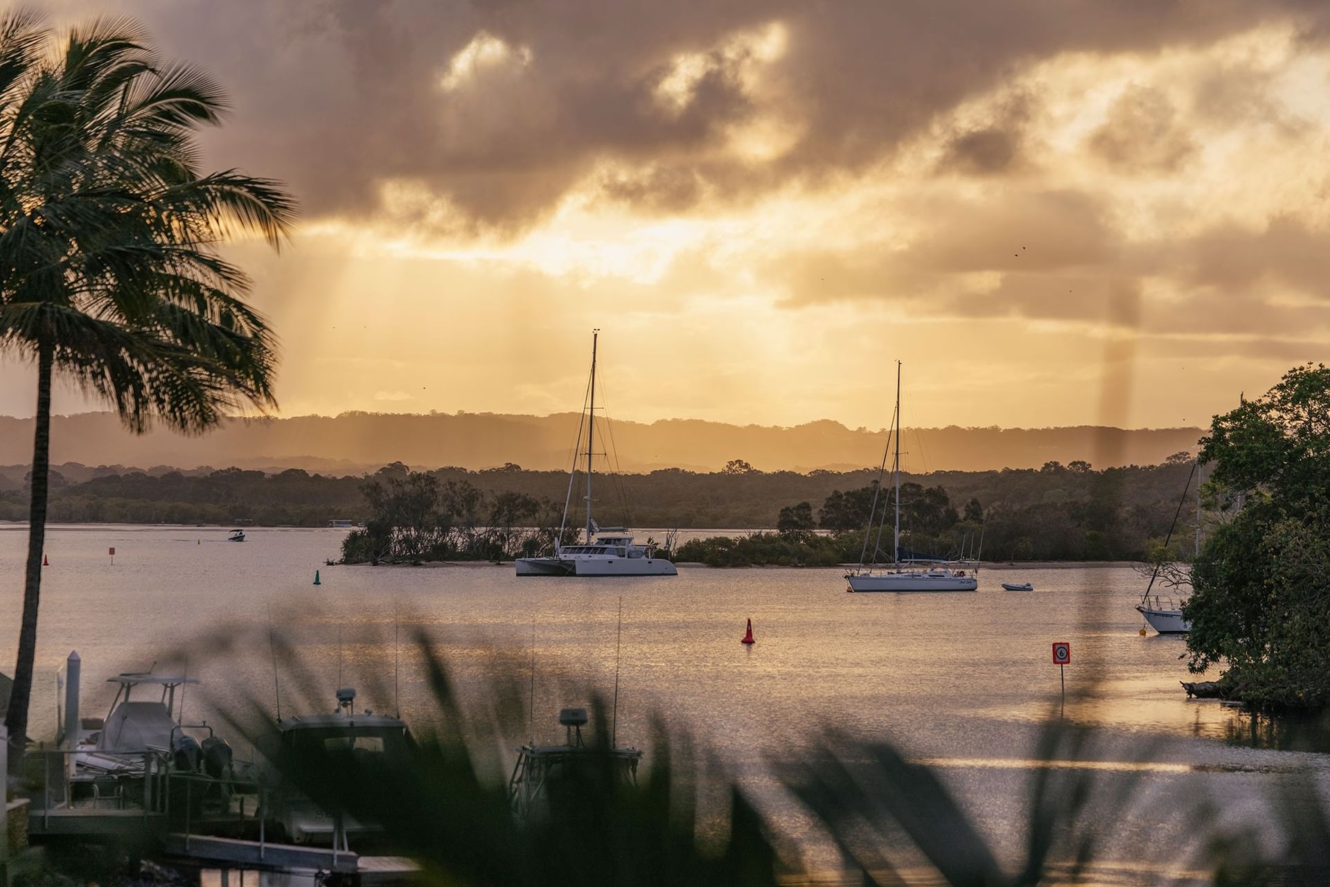 Sun setting over the Noosa River at Elysium Noosa Resort with sailboats floating against a warm golden sunset over lush hills
