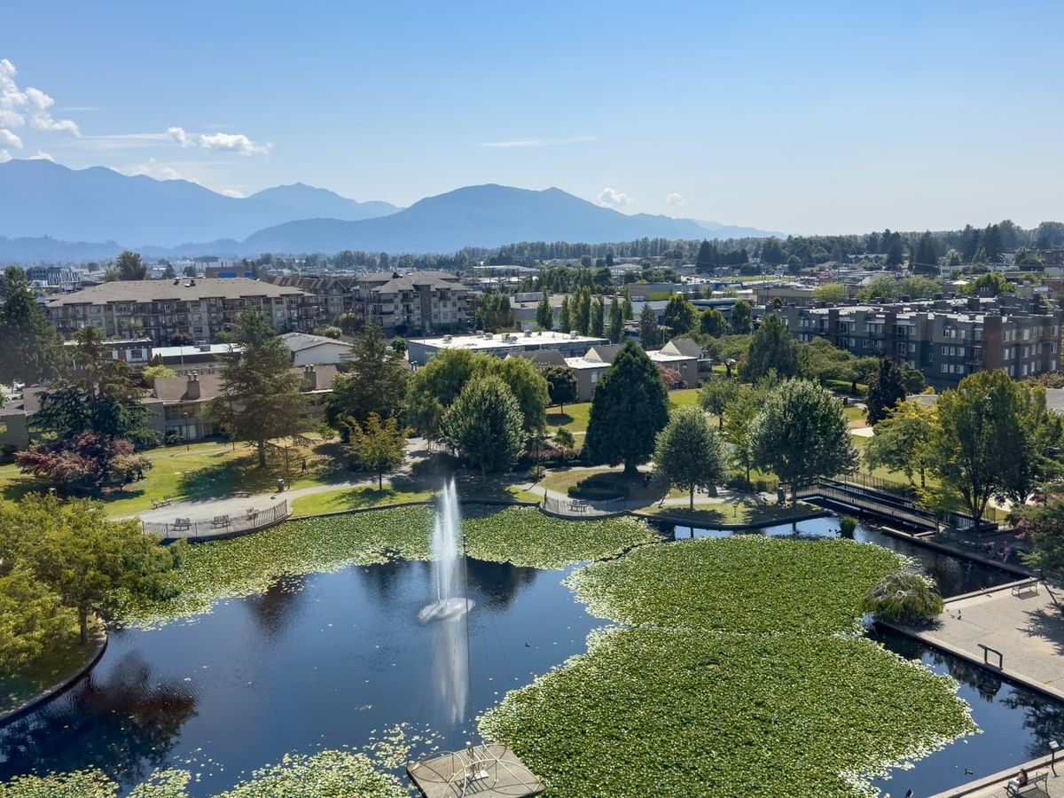 Aerial view of a pond with a fountain, surrounded by greenery, buildings, and distant mountains.