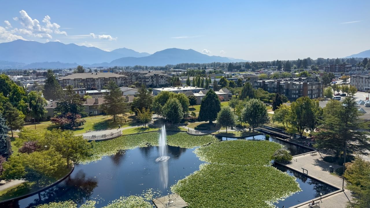 Aerial view of a pond with a fountain, surrounded by greenery, buildings, and distant mountains.