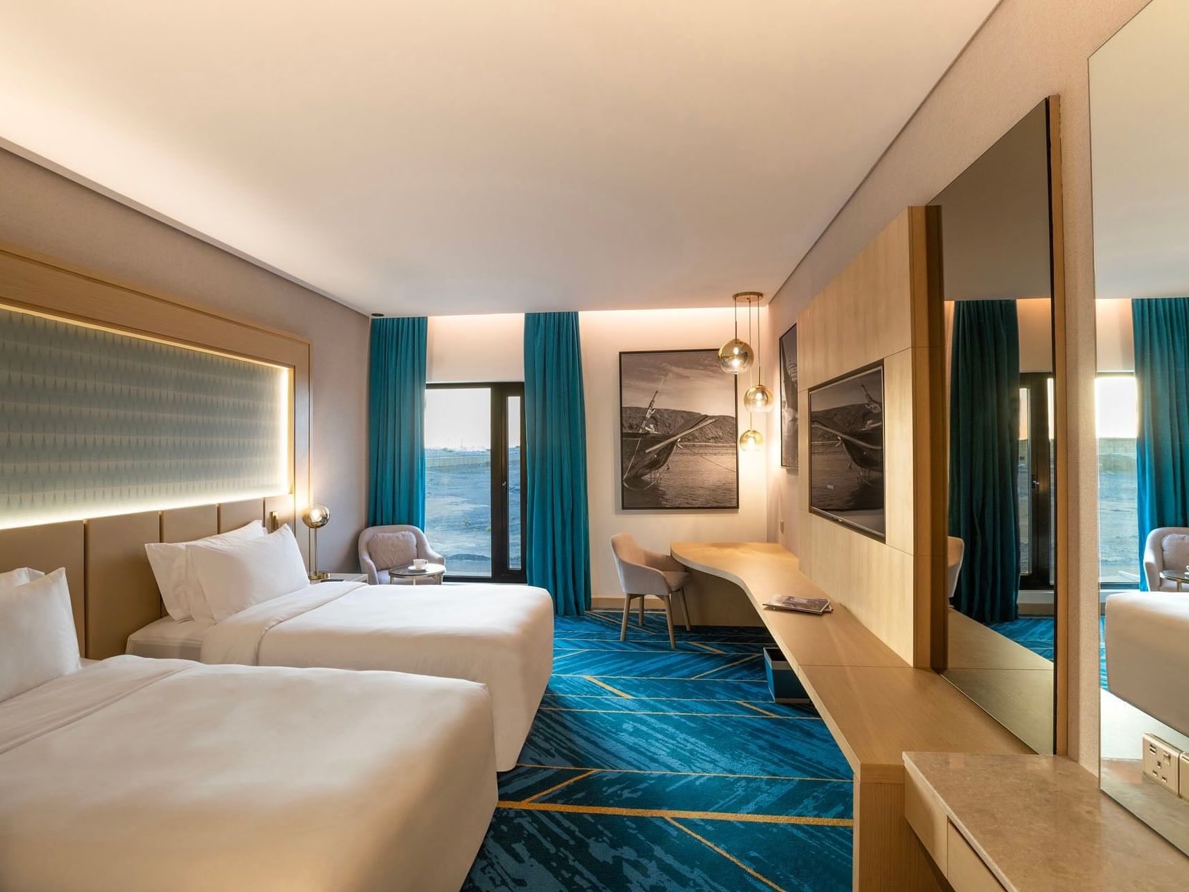 A hotel room with two beds, a desk, a chair, and a balcony with blue curtains.