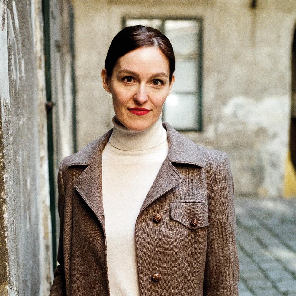 A woman in a white turtleneck and brown jacket stands against a wall near a building.