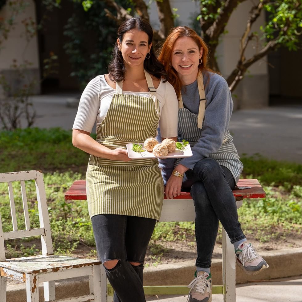 Two smiling chefs in striped aprons presenting plant-based dishes on white plates in a sunny garden setting.