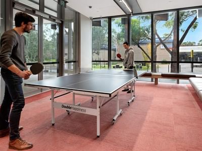 Two men playing table tennis in a room with large windows and a couch at La Trobe University.