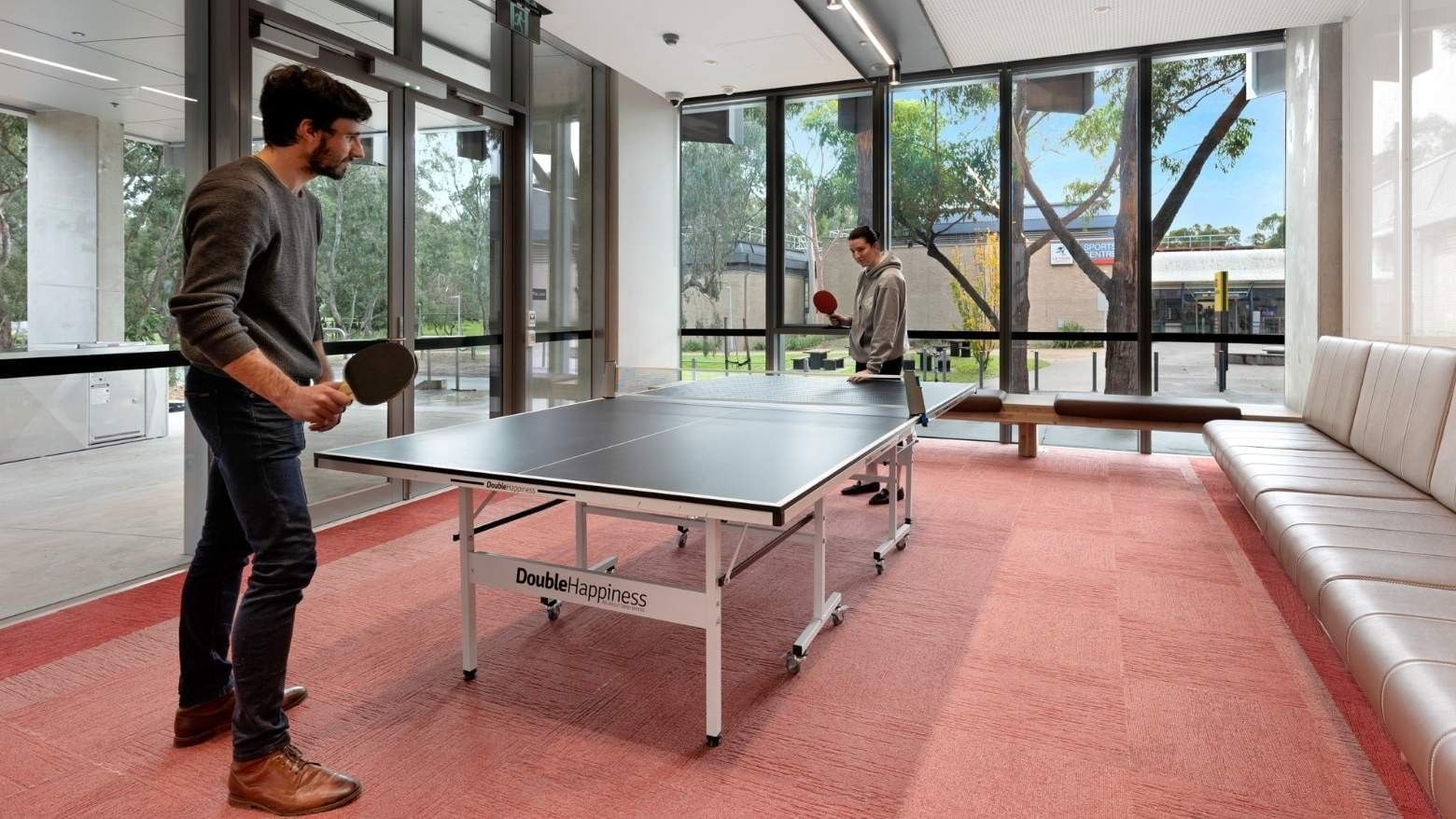 Two men playing table tennis in a room with large windows and a couch at La Trobe University.