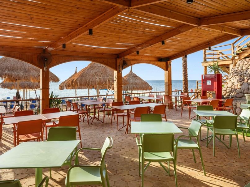 Outdoor restaurant with wooden roof, green and orange tables and chairs, and thatched umbrellas by the beach.