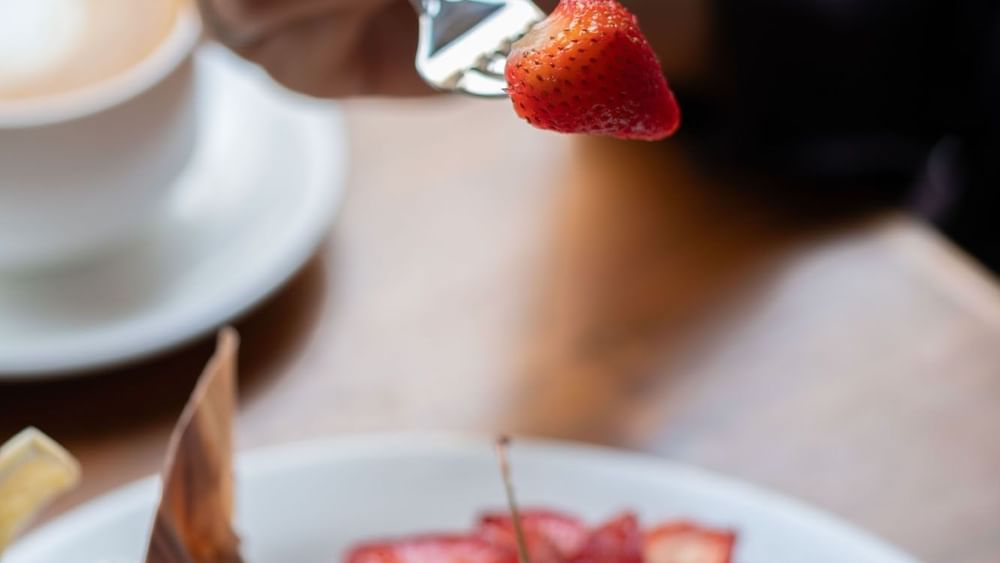 Hand holding a fork with a strawberry over a plate of cheesecake in Artist Lobby Lounge at Warwick Hotel Jeddah