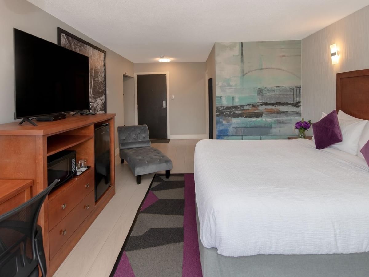 A hotel room with a bed, purple pillows, a TV, a desk, a chair, and a rug.