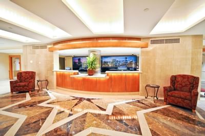 A luxurious lobby with a marble floor, two monitors, two armchairs, and a reception desk with a plant.
