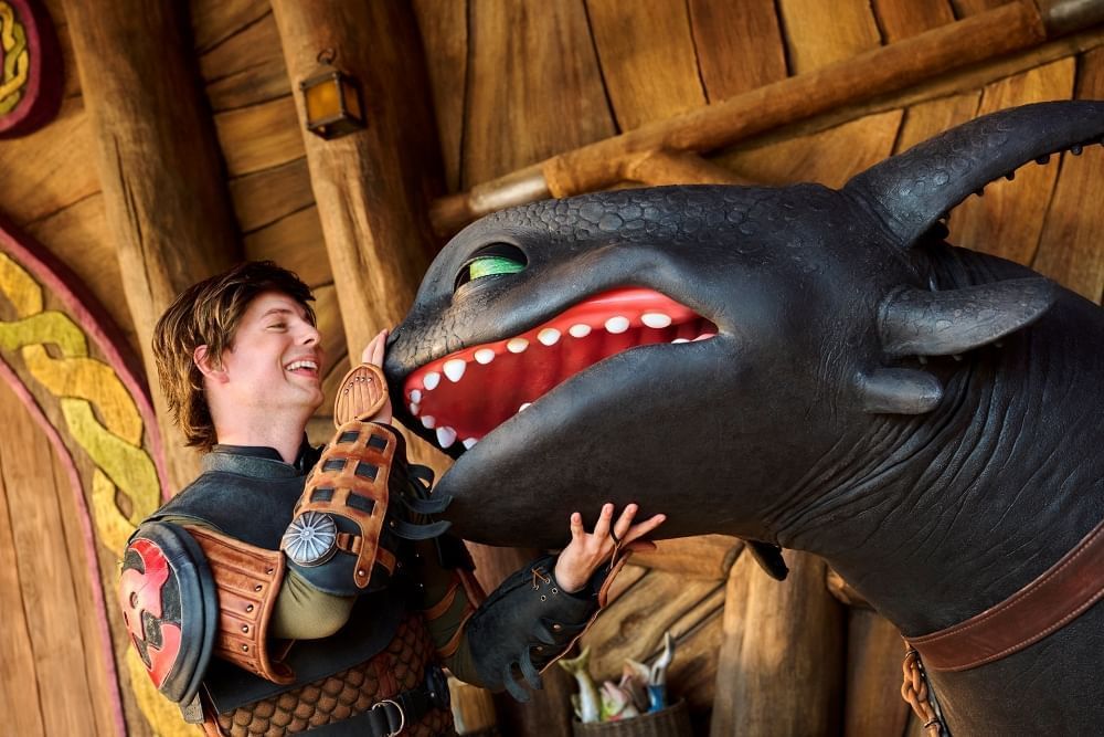 A young brunette man in Viking attire pets the nose of a black, smiling dragon.