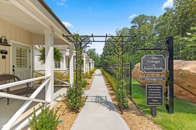 Sunny walkway lined with greenery leads to The Cottages at Laurel Brooke, topped by a welcoming sign and clear blue sky