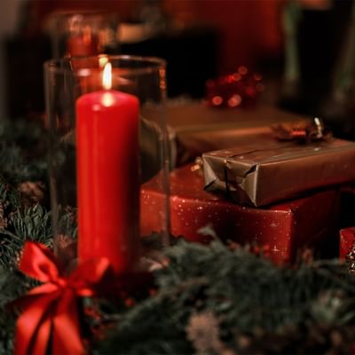 Rote Kerze in Glas neben Geschenken und grünem Kranz mit rotem Band.