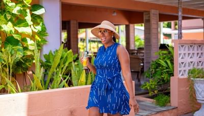 Happy woman in blue dress and sun hat holding a yellow cocktail drink by green plants at Worthing Court Apartment