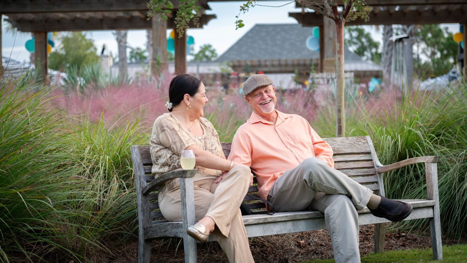 Senior couple relaxing on a wooden bench in a landscaped garden area with pink ornamental grasses and pergola.