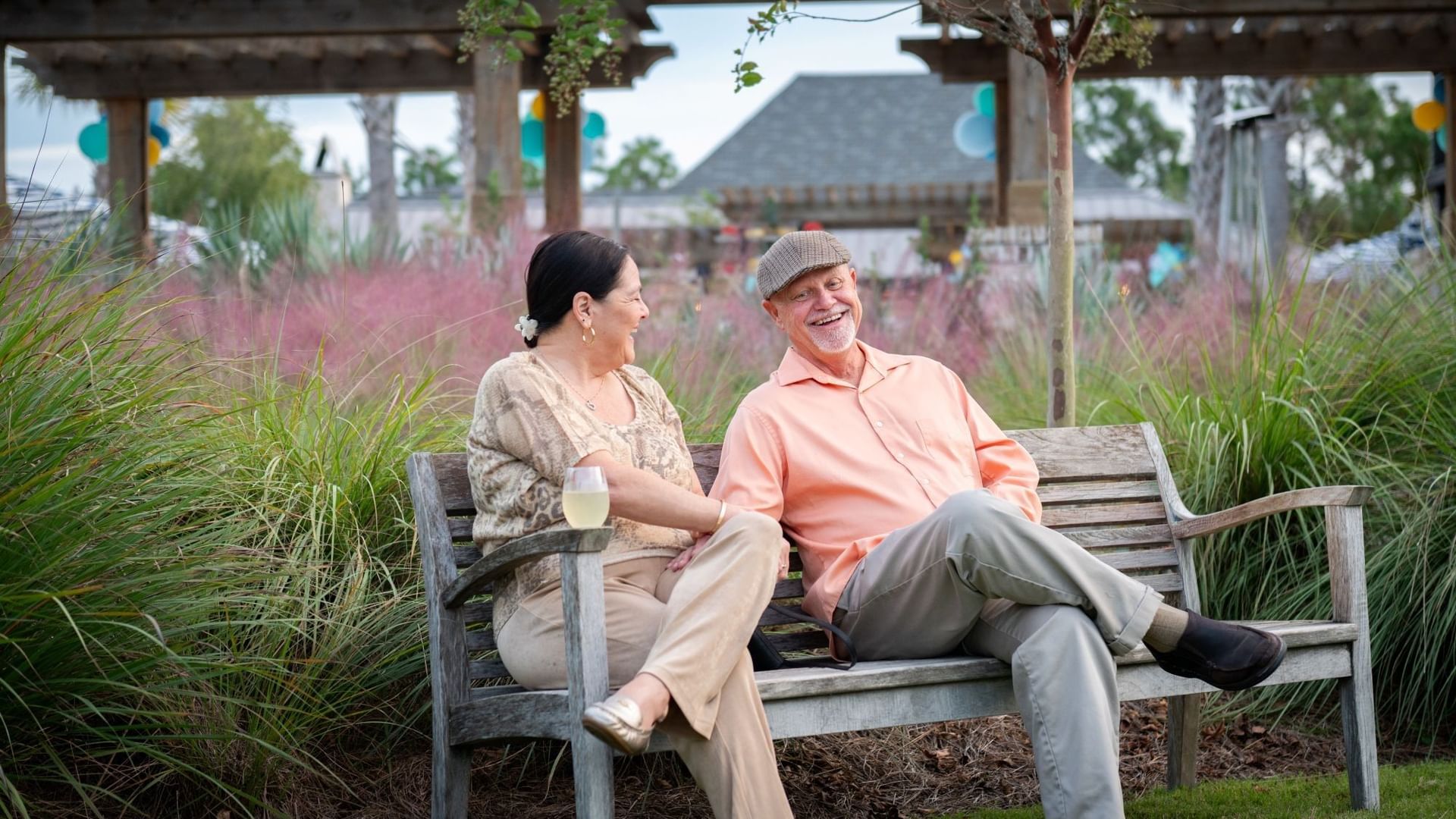 Senior couple relaxing on a wooden bench in a landscaped garden area with pink ornamental grasses and pergola.