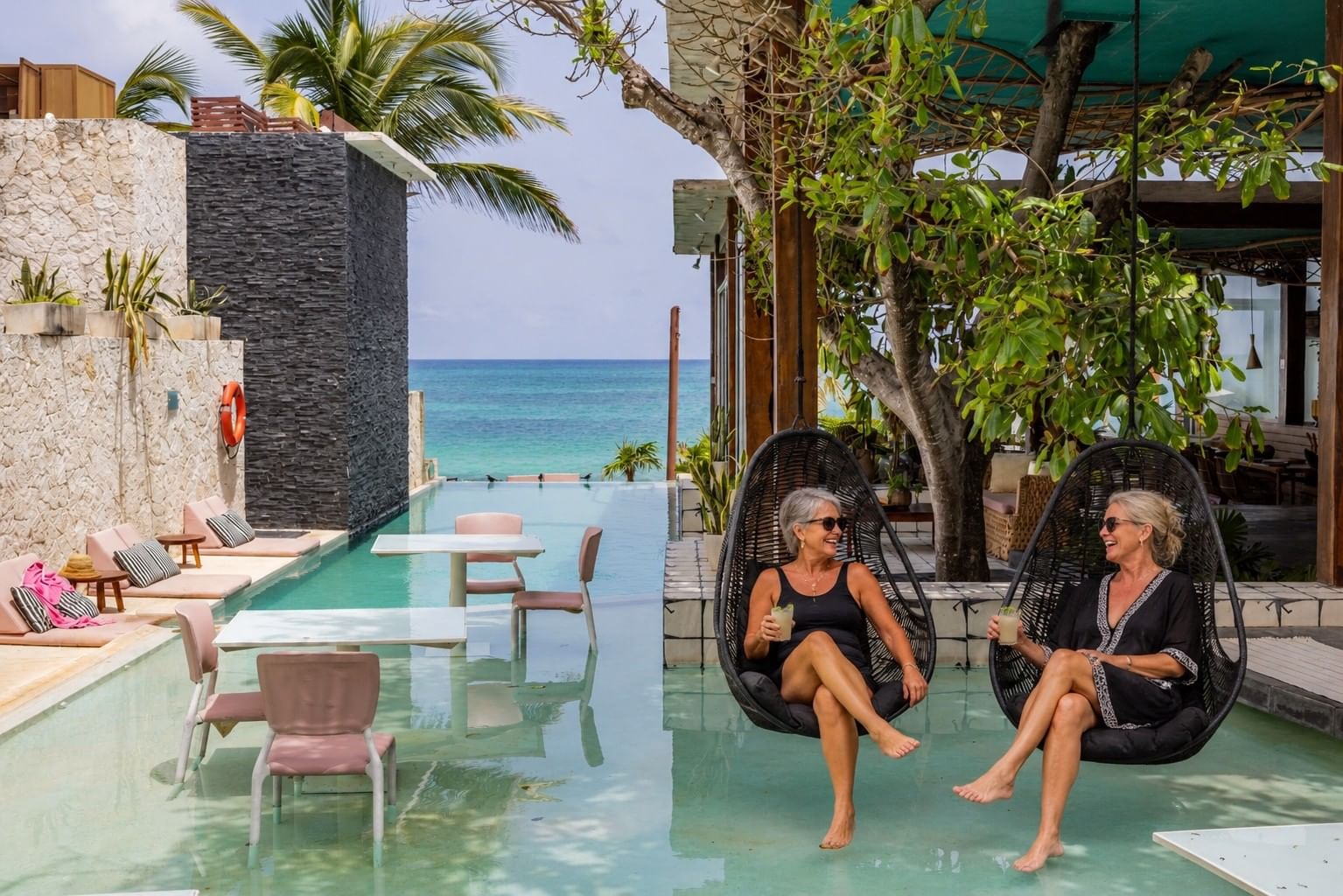 Two women enjoying drinks in hanging chairs beside Mi Amor's infinity pool