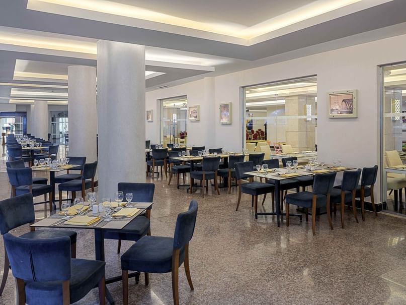 An empty restaurant with blue chairs, rectangular tables, and glass walls showing the outdoors.