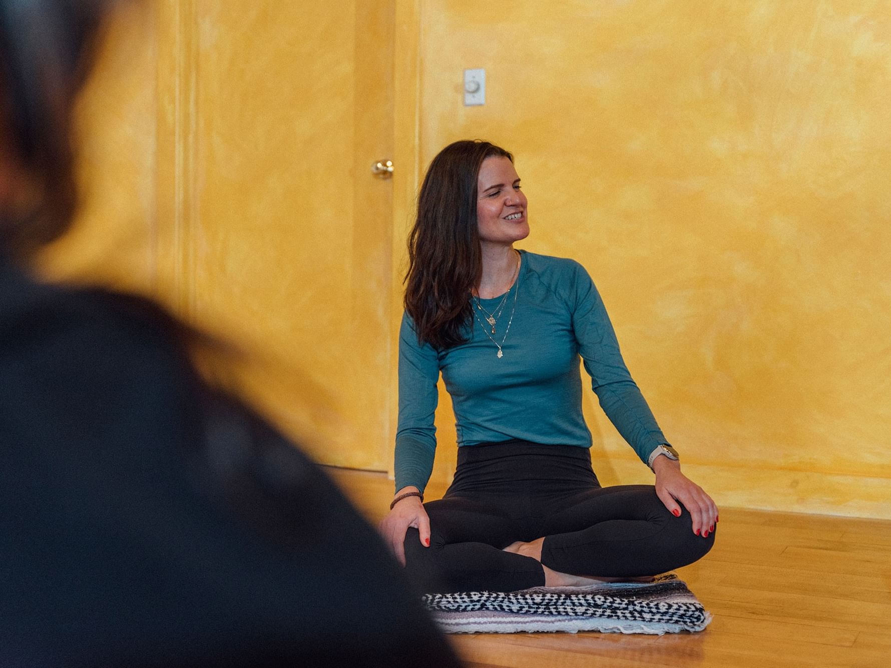 Woman in teal shirt and black pants sitting in lotus position on patterned mat against yellow wall.