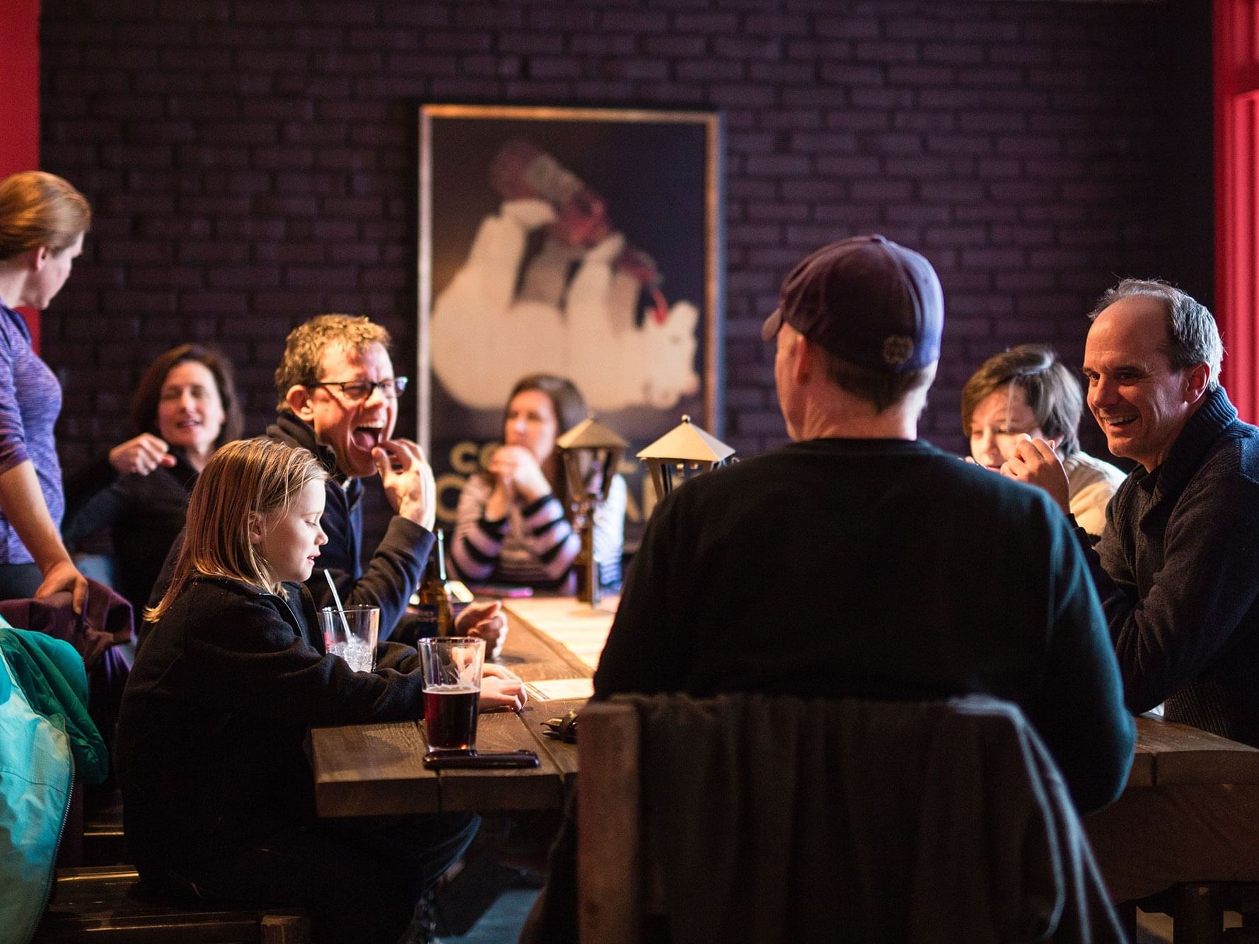 Group of guests enjoying conversation and drinks together at a communal dining table in a warmly lit restaurant setting.