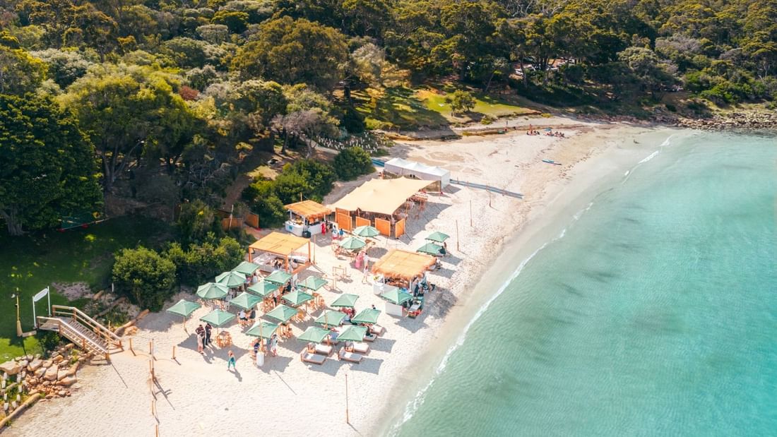 Aerial view of a beachside event setup with turquoise umbrellas and seating on white sand surrounded by lush greenery.