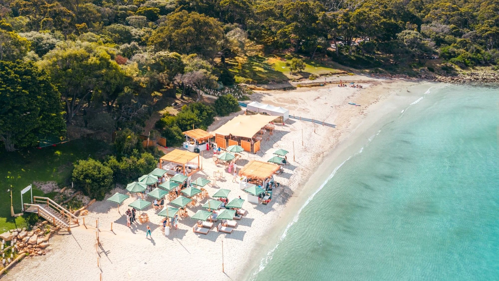 Aerial view of a beachside event setup with turquoise umbrellas and seating on white sand surrounded by lush greenery.