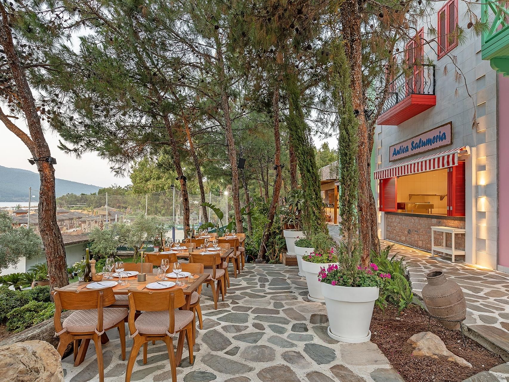 An outdoor restaurant at Titanic Luxury Collection Bodrum, set on a stone floor with tables, chairs, flower pots, and a sea view in the background.