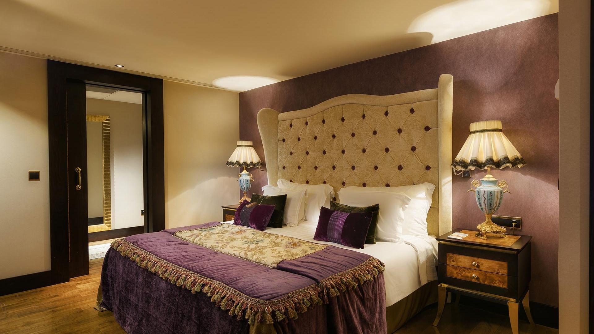 Elegant bedroom with large bed, purple bedding, wooden floors, and ornate nightstands with lamps in Pool Duplex Suite at Titanic Mardan Palace.