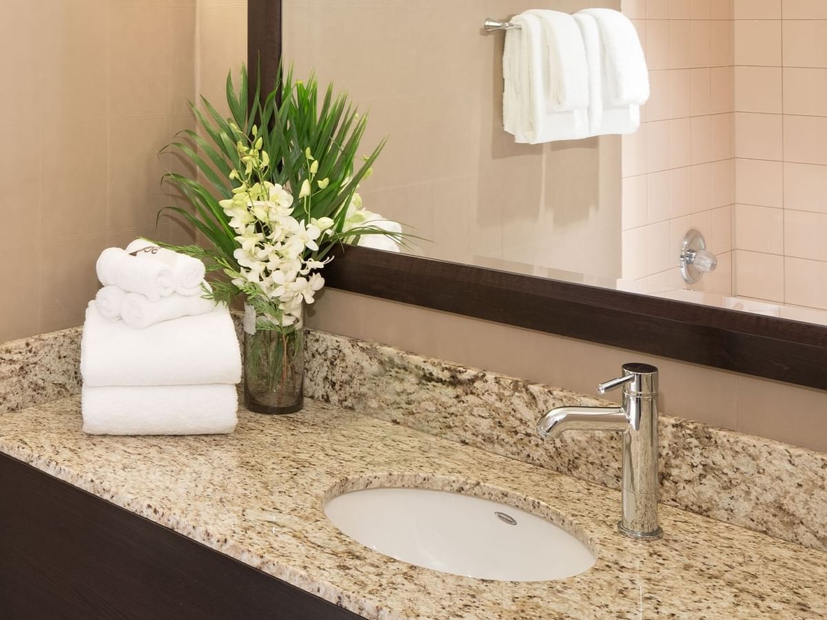 A bathroom vanity with a sink, faucet, towel, and flowers in a vase, with a mirror reflecting a shower.