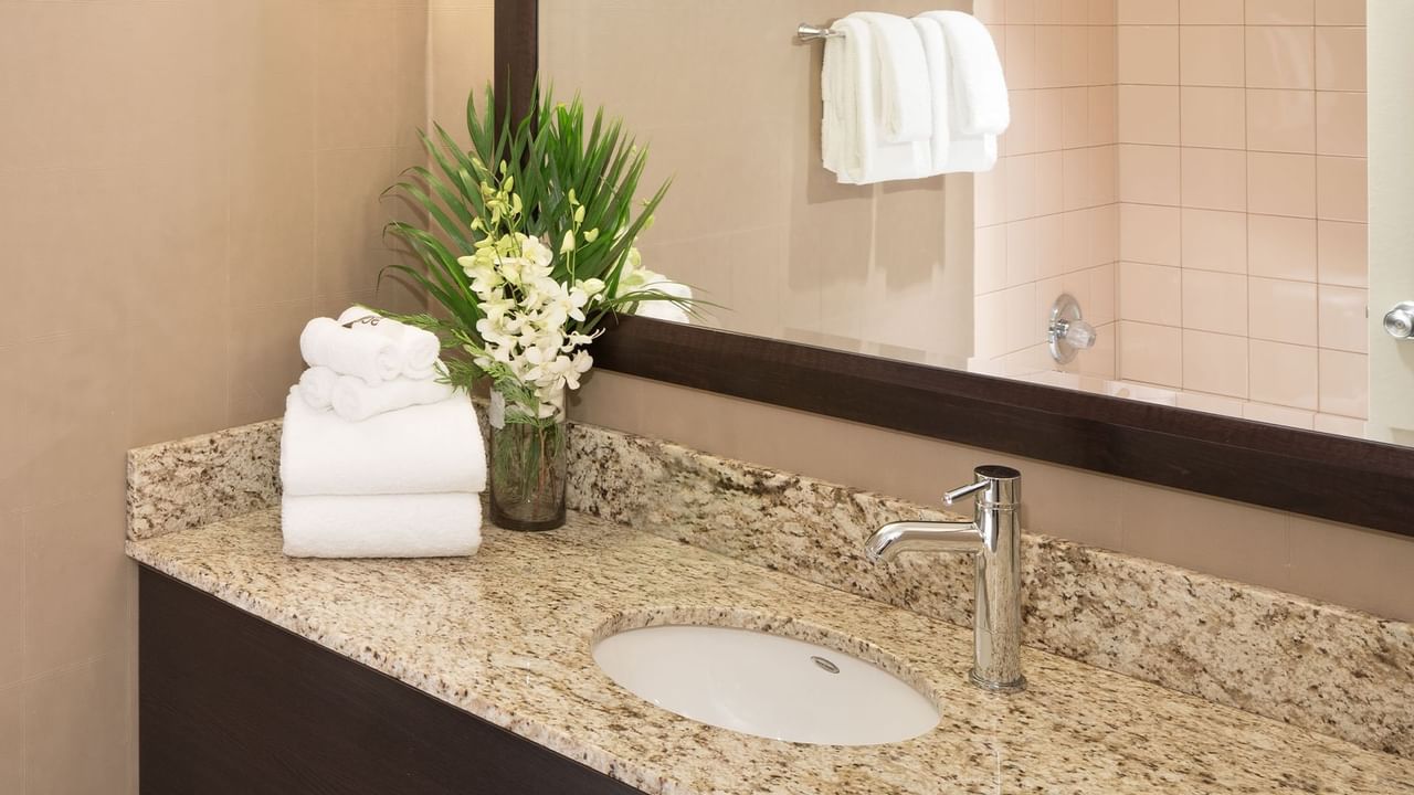 A bathroom vanity with a sink, faucet, towel, and flowers in a vase, with a mirror reflecting a shower.