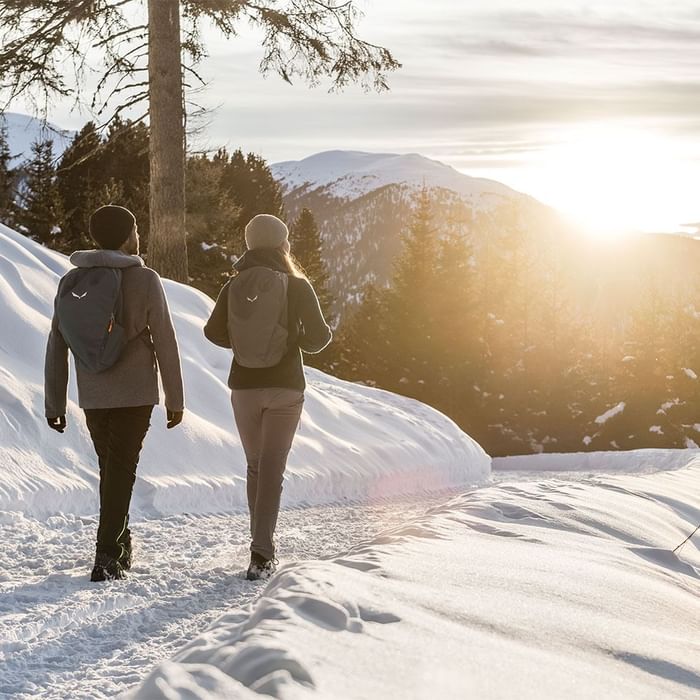 Two people hiking in snow towards the sunset among mountains near Falkensteiner Hotel Antholz