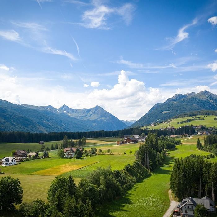 Panorama di una vallata con case, prati, alberi e montagne sullo sfondo sotto un cielo blu.