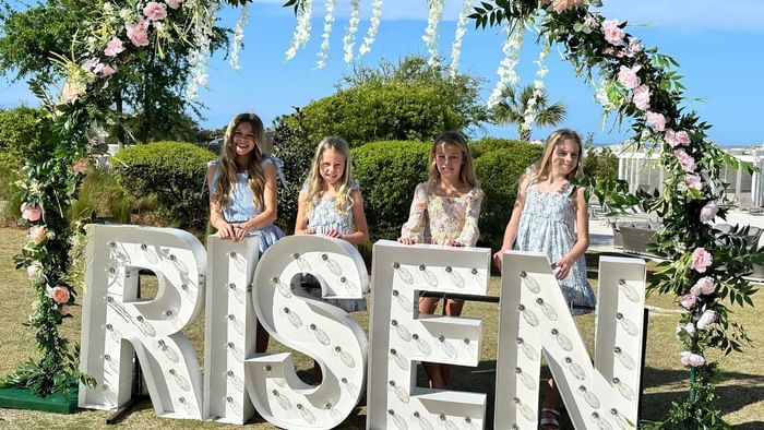 Four girls stand behind a decorated arch and large sign spelling RISEN for Easter Weekend on 30A.